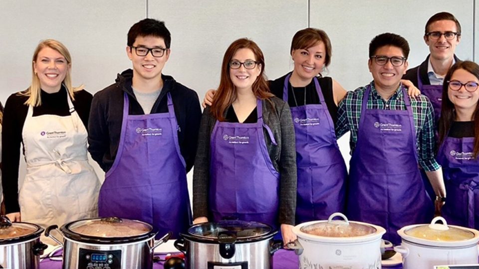 Eight volunteers wearing aprons, standing behind a table with food cooking.