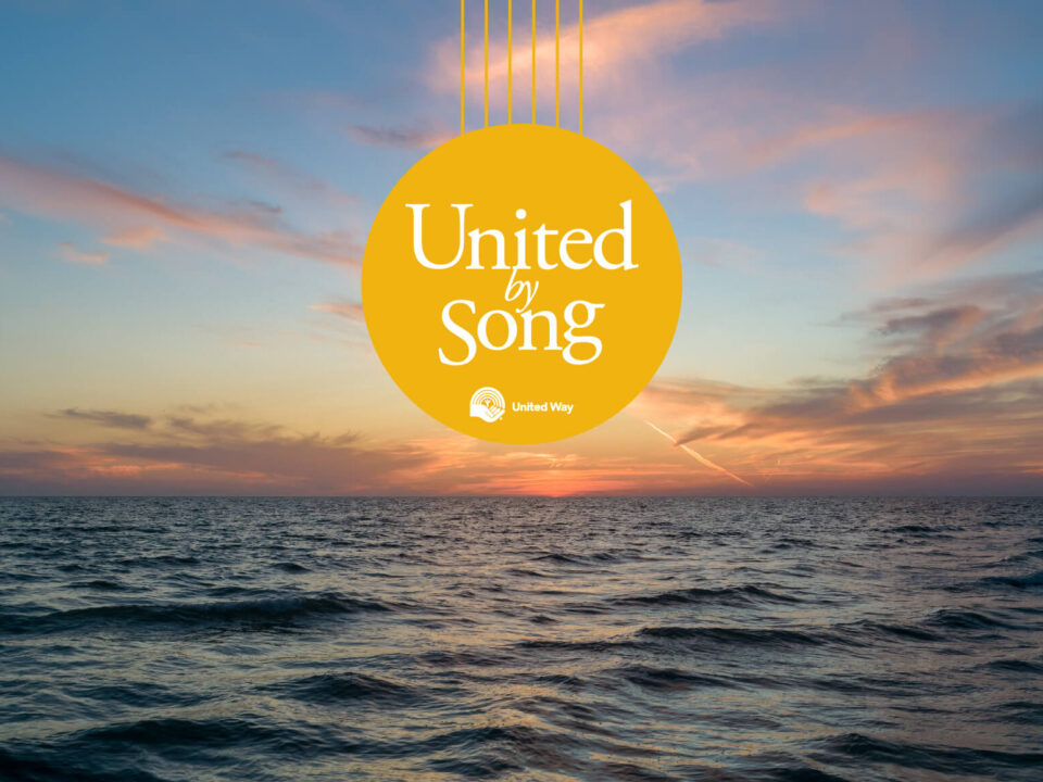 Sunset over open ocean with thin, pink clouds. In the centre of the image is a yellow circle with white text reading, United by Song.