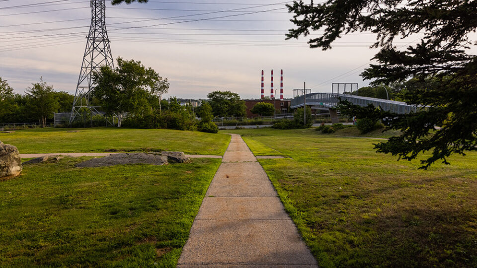 A concrete path in Dartmouth North with green grass on either side and red and white smoke stacks in the distance