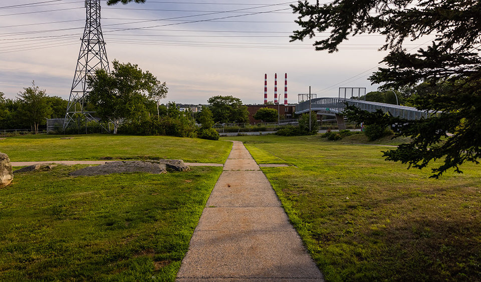 A concrete path in Dartmouth North with green grass on either side and red and white smoke stacks in the distance