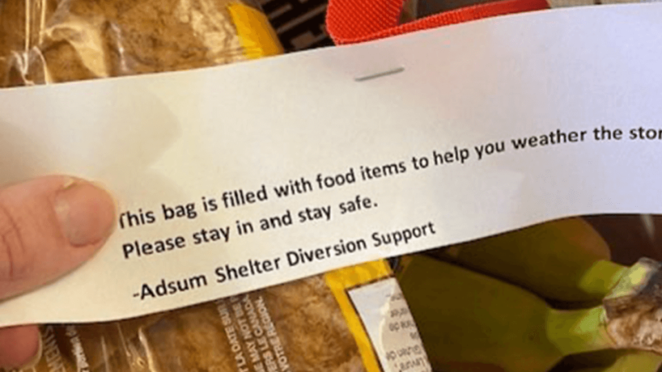 A grocery bag with bread, fruit and other items. A hand holds a note to accompany it: This bag is filled with food items to help you weather the storm. Please stay in and stay safe. Adsum Shelter Diversion Support