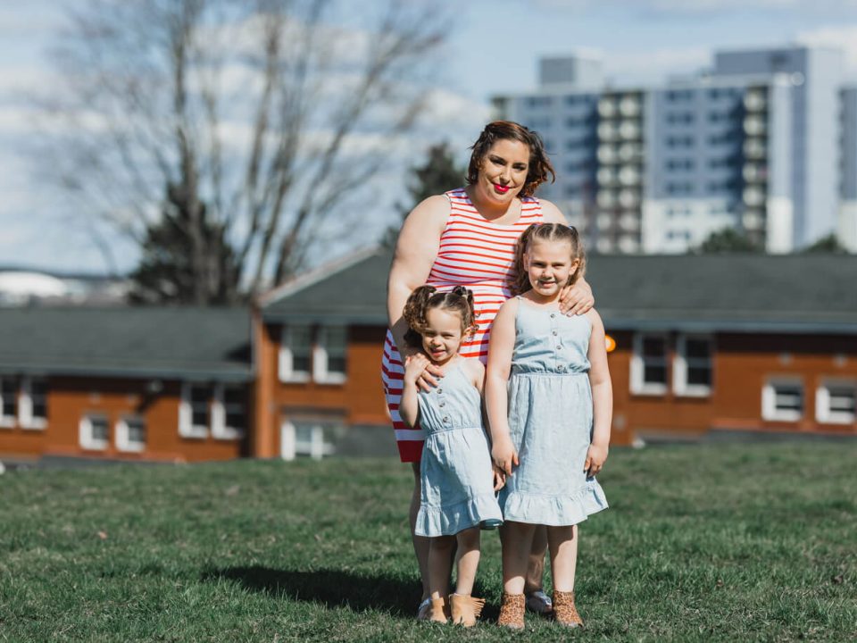 A woman in a red striped dress standing behind her two daughters in baby blue dresses. They're standing in an open grassy area with houses visible behind her. She accesses the Lived Experience Honorarium.