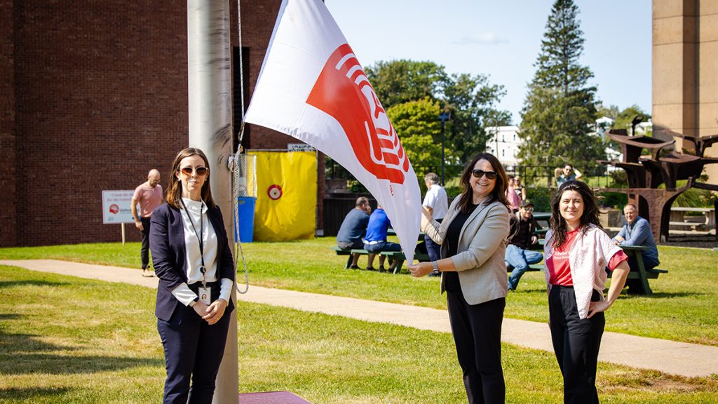 Three woman standing near a flagpole with the woman in the middle holding a United Way flag
